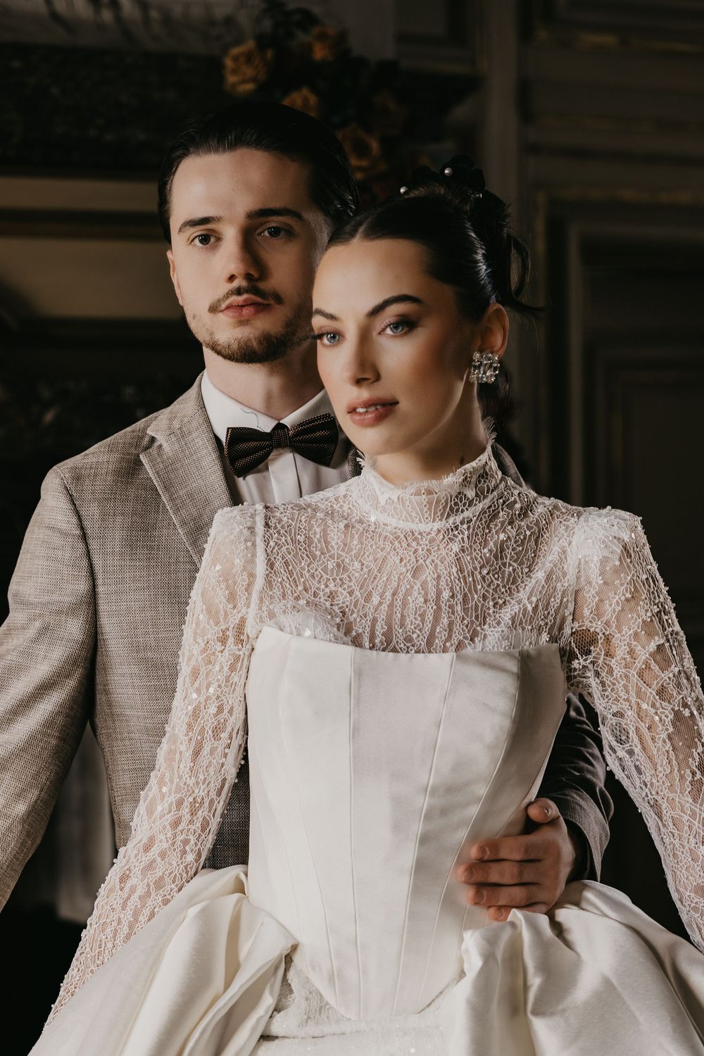 A couple in elegant wedding attire poses together, with the bride wearing a vintage-style lace gown with high neckline.