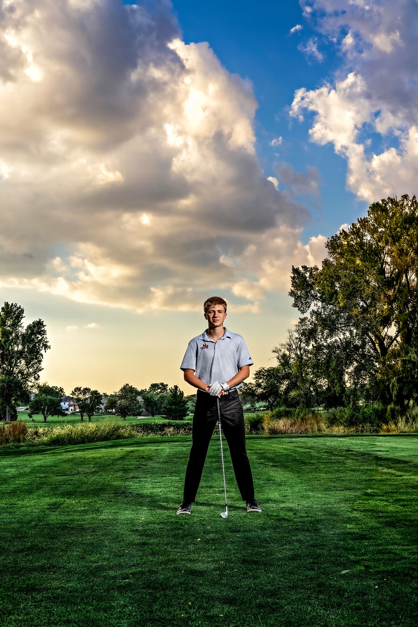 A golfer stands on a lush green course at sunset with dramatic clouds and trees in the background.