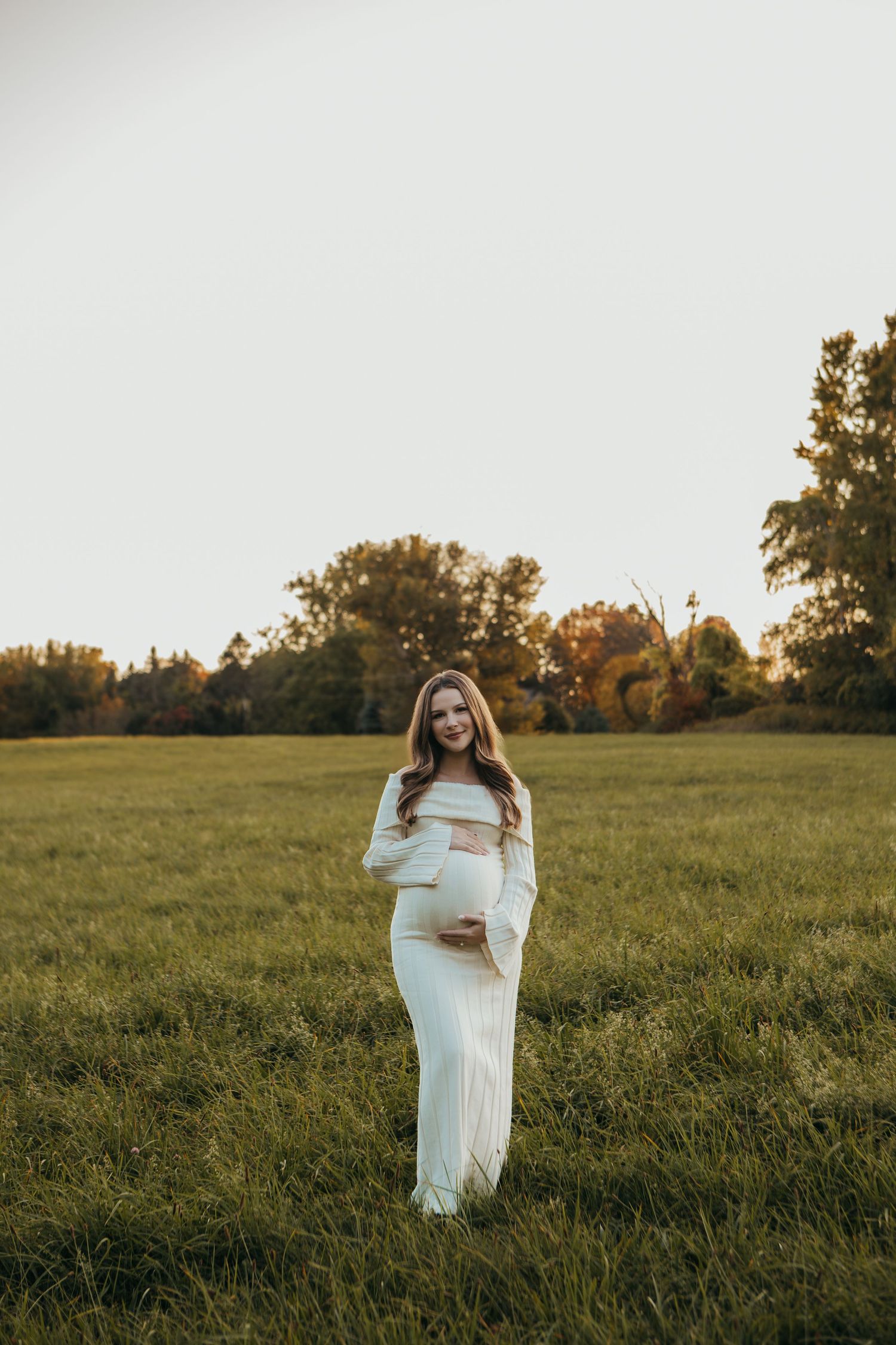 Maternity photoshoot in a sunlit field with flowing white dress at golden hour.