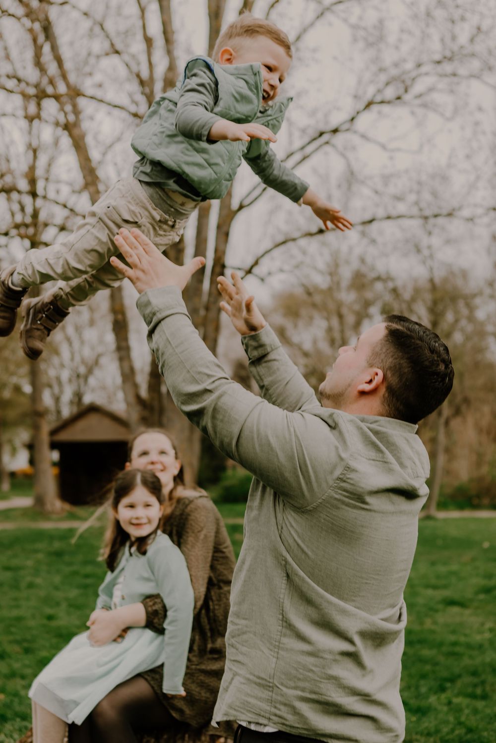 A man playfully throws a laughing young boy into the air while a woman and a young girl watch and smile from the background in a park.