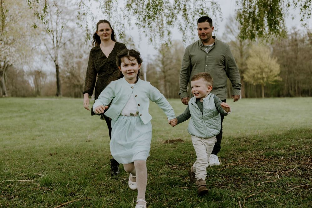 A smiling young girl and boy run hand-in-hand through a grassy field, followed closely by a man and woman walking behind them in a park.