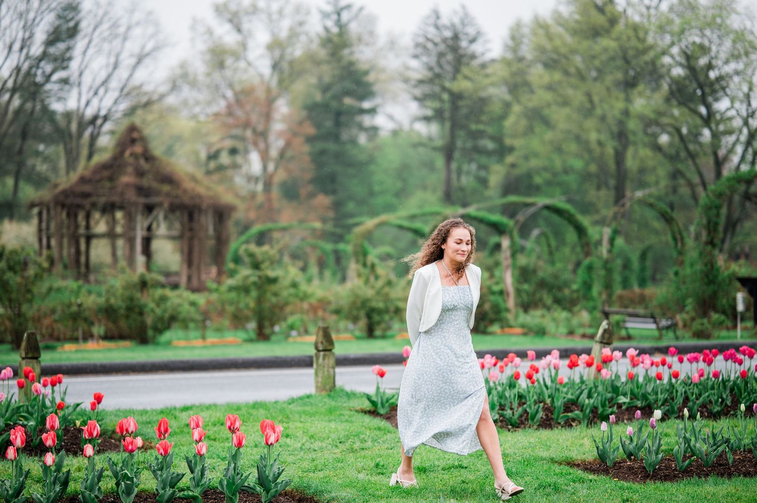 A white dress flows in a spring garden with vibrant pink tulips and a rustic gazebo in the background.