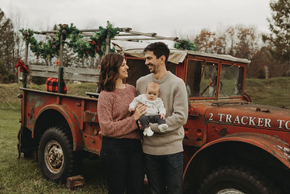 A couple stands with a baby in front of a vintage red tractor during an autumn photoshoot at a farm.
