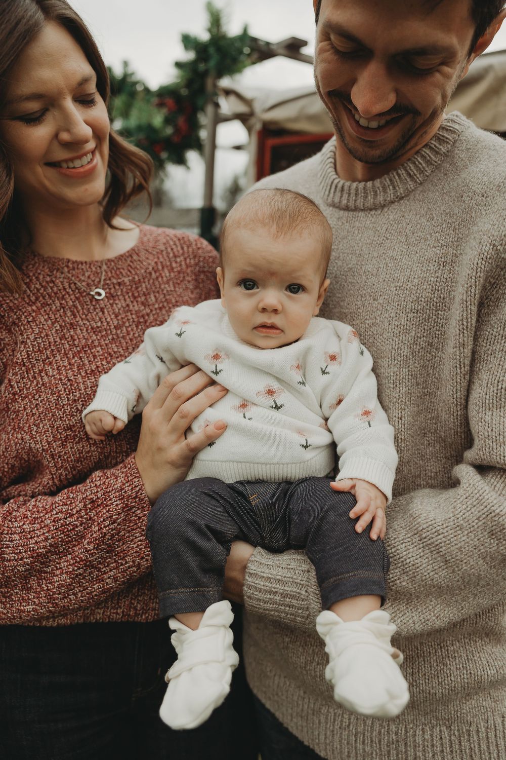 A couple in knit sweaters holding their baby wearing a white patterned outfit and white booties.