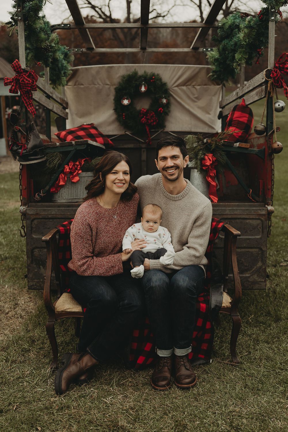 Family holiday portrait on rustic bench decorated with Christmas wreaths and red buffalo plaid accents.