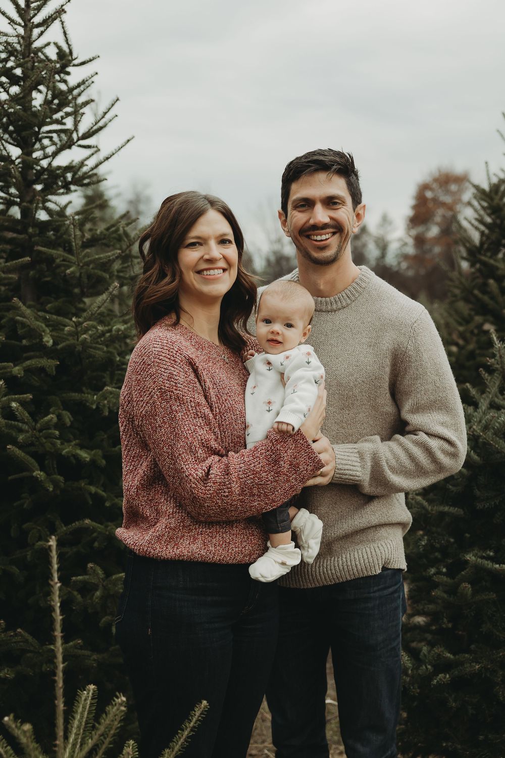 Family portrait at Christmas tree farm with parents in sweaters holding their baby against evergreen backdrop.