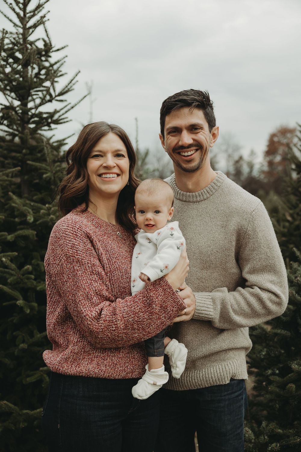 Family wearing cozy sweaters poses together for holiday photos among evergreen trees on an overcast day.