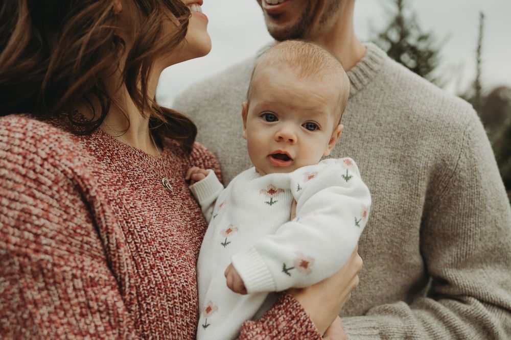A couple in cozy autumn sweaters holds a baby in a white onesie during an outdoor family photo session.