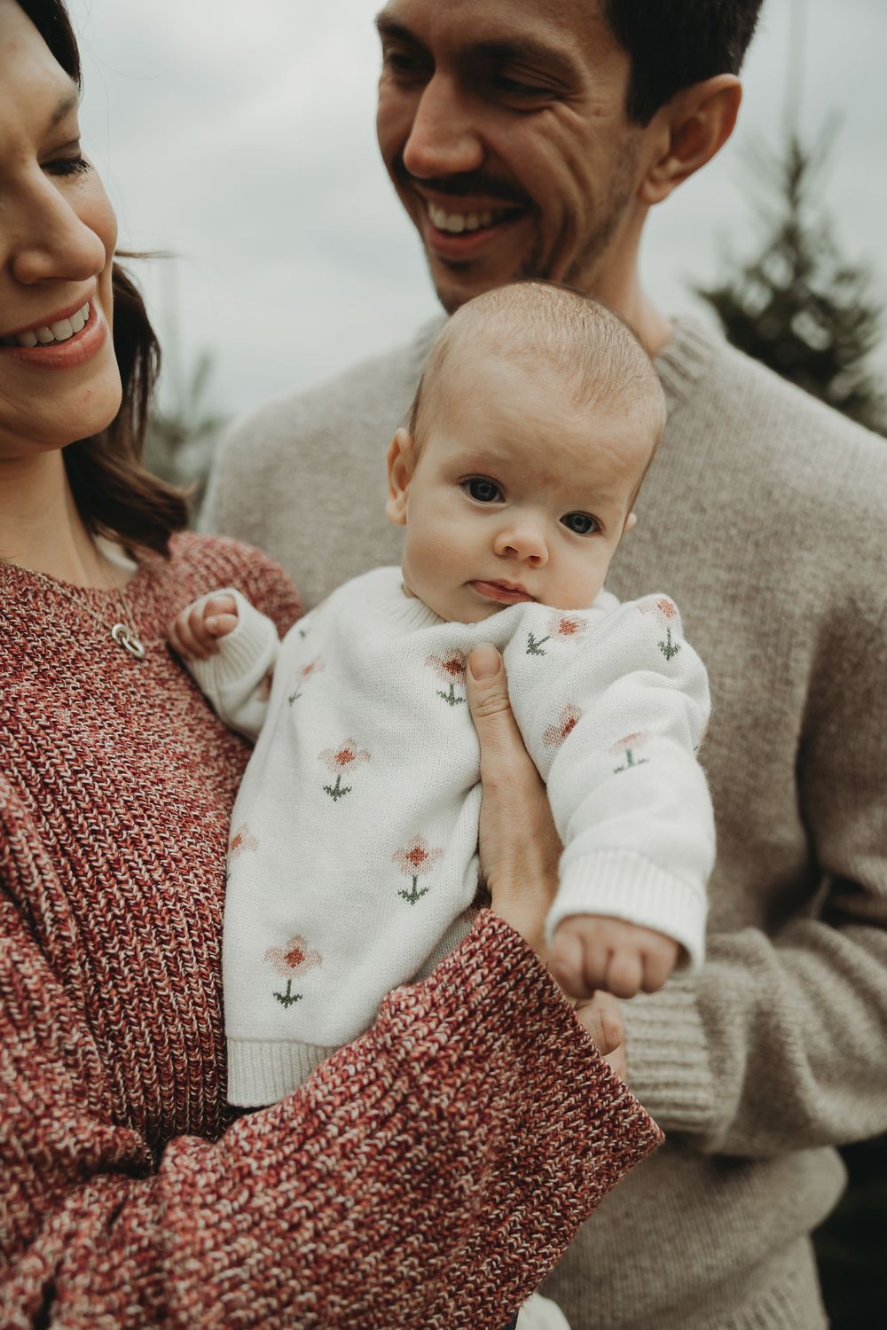 A baby wearing a white onesie with bunny face design is held by adults in casual winter clothing.