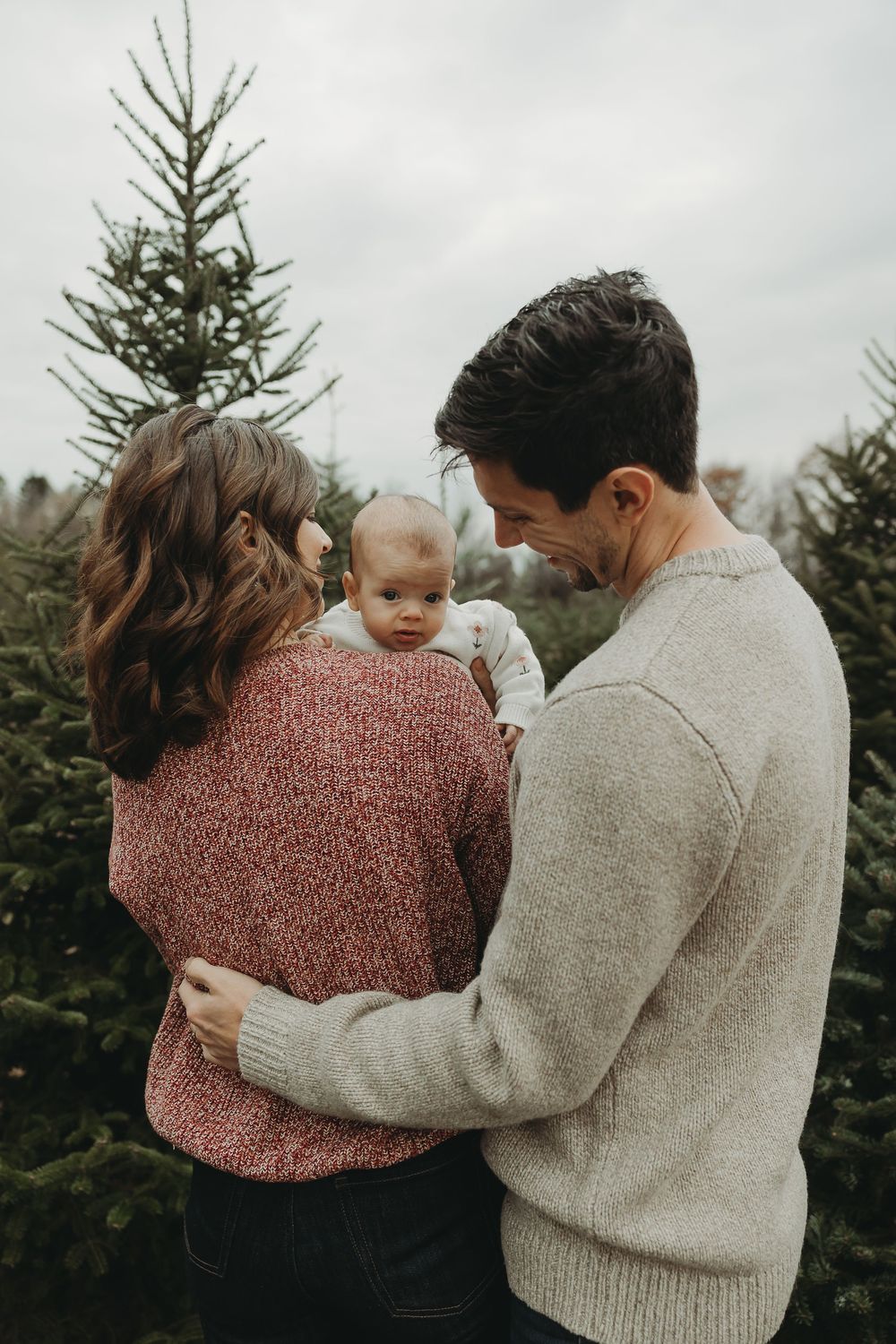 Family in cozy sweaters sharing tender moments together at a Christmas tree farm on an overcast day.