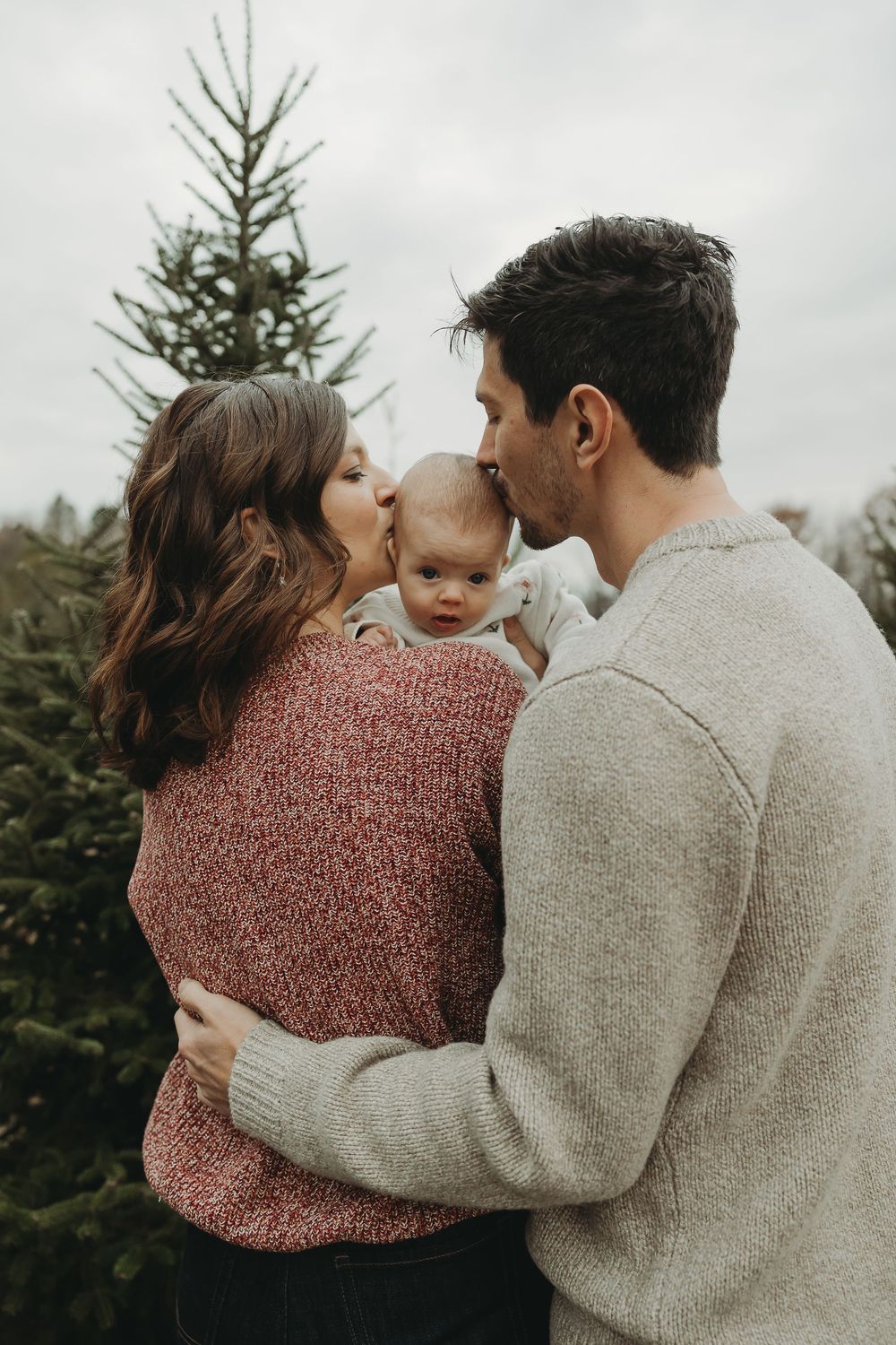 Intimate family portrait showing loving embraces between parents and baby among evergreen trees.