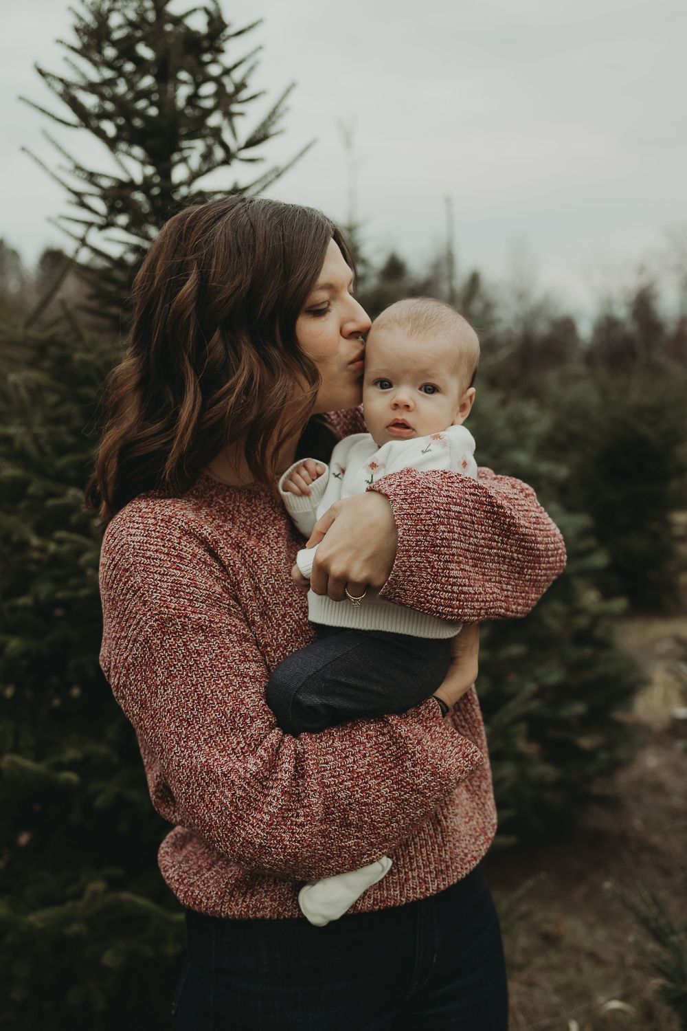 A tender moment between parent and baby among evergreen trees in a moody winter setting.