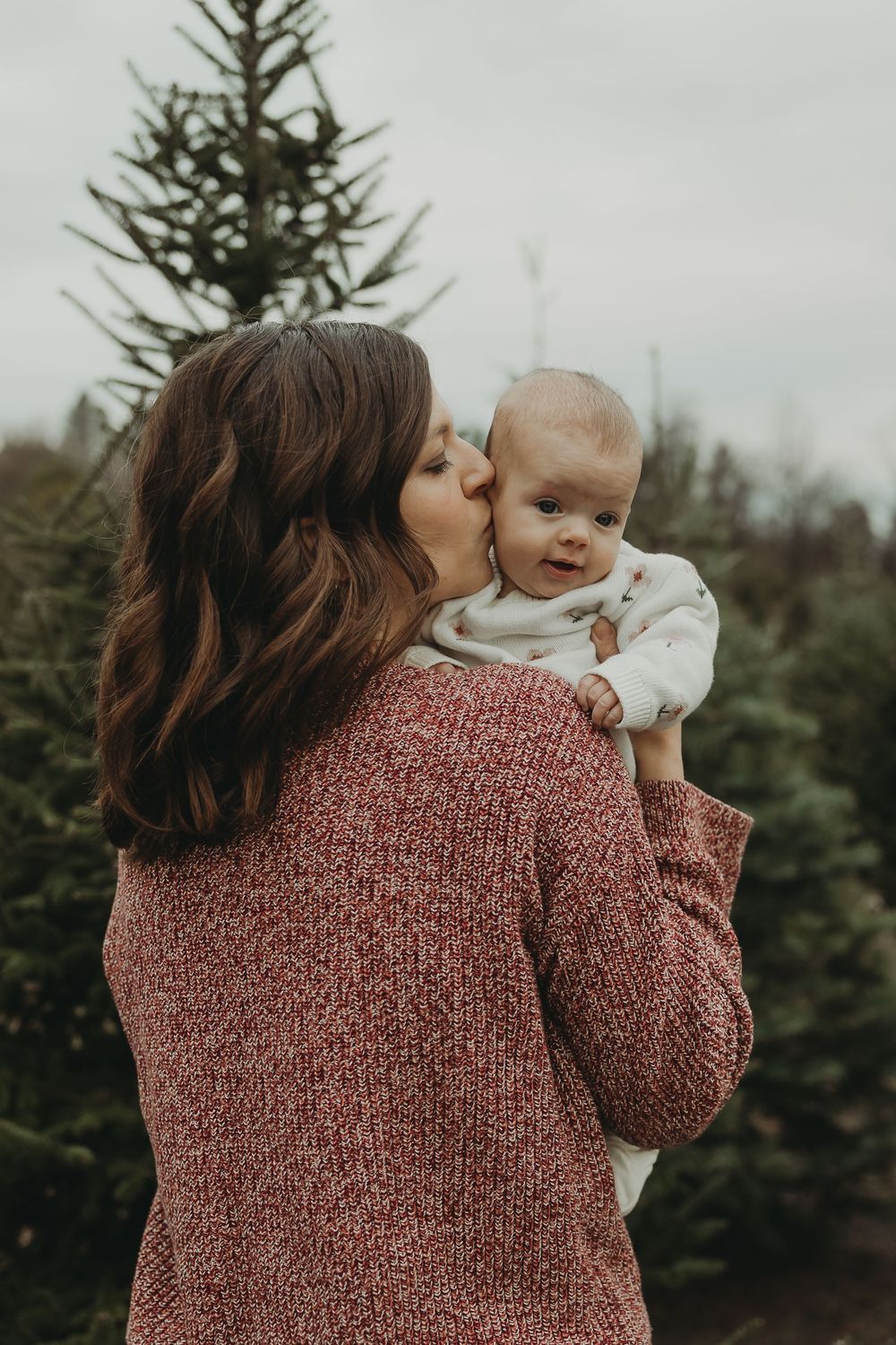 A series of tender moments captured between a parent and baby at a Christmas tree farm in matching burgundy sweaters.