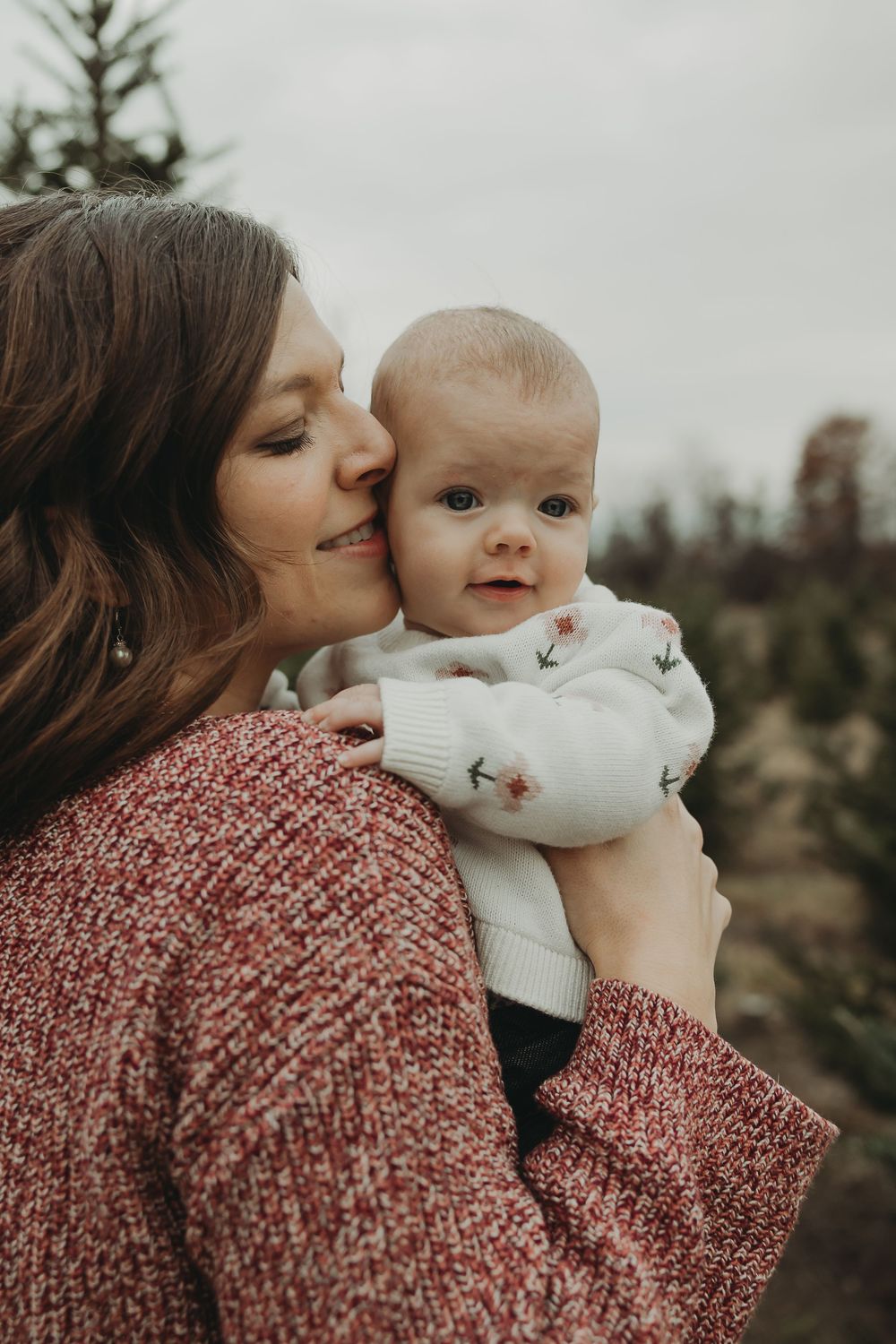 A tender moment captured outdoors as a mother in a red sweater holds her baby wearing white against a moody sky.