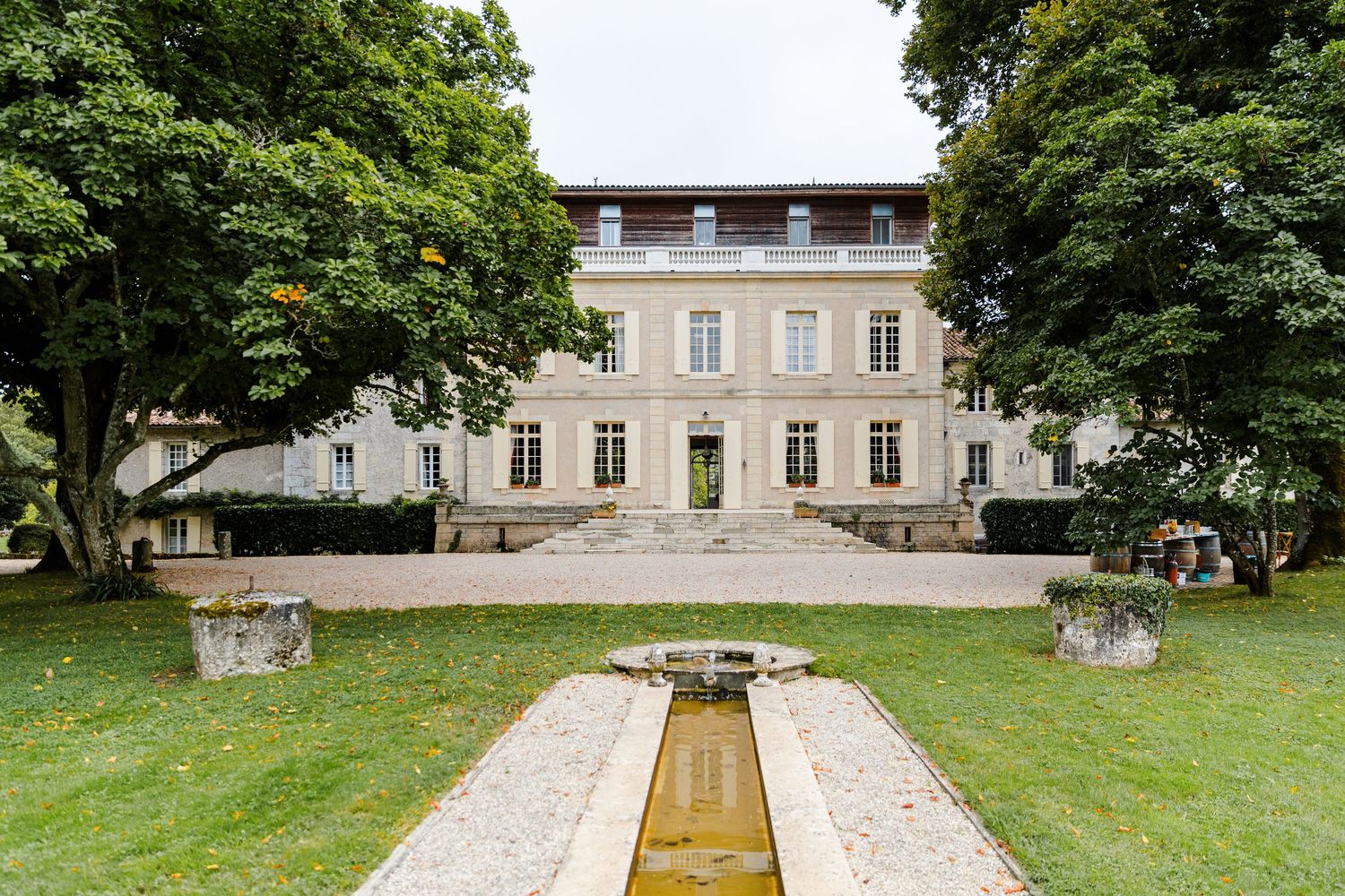 A grand estate garden features a long stone pathway leading to a historic mansion through manicured lawns and trees.