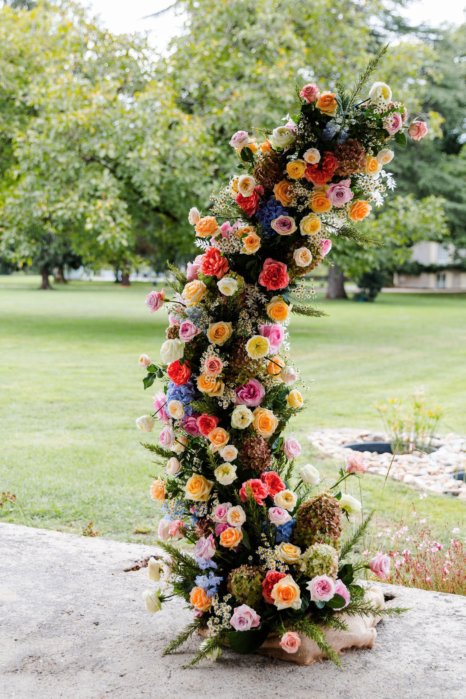 A vibrant floral arrangement with pink, orange, and red roses cascades down a stone wall in an outdoor garden setting.
