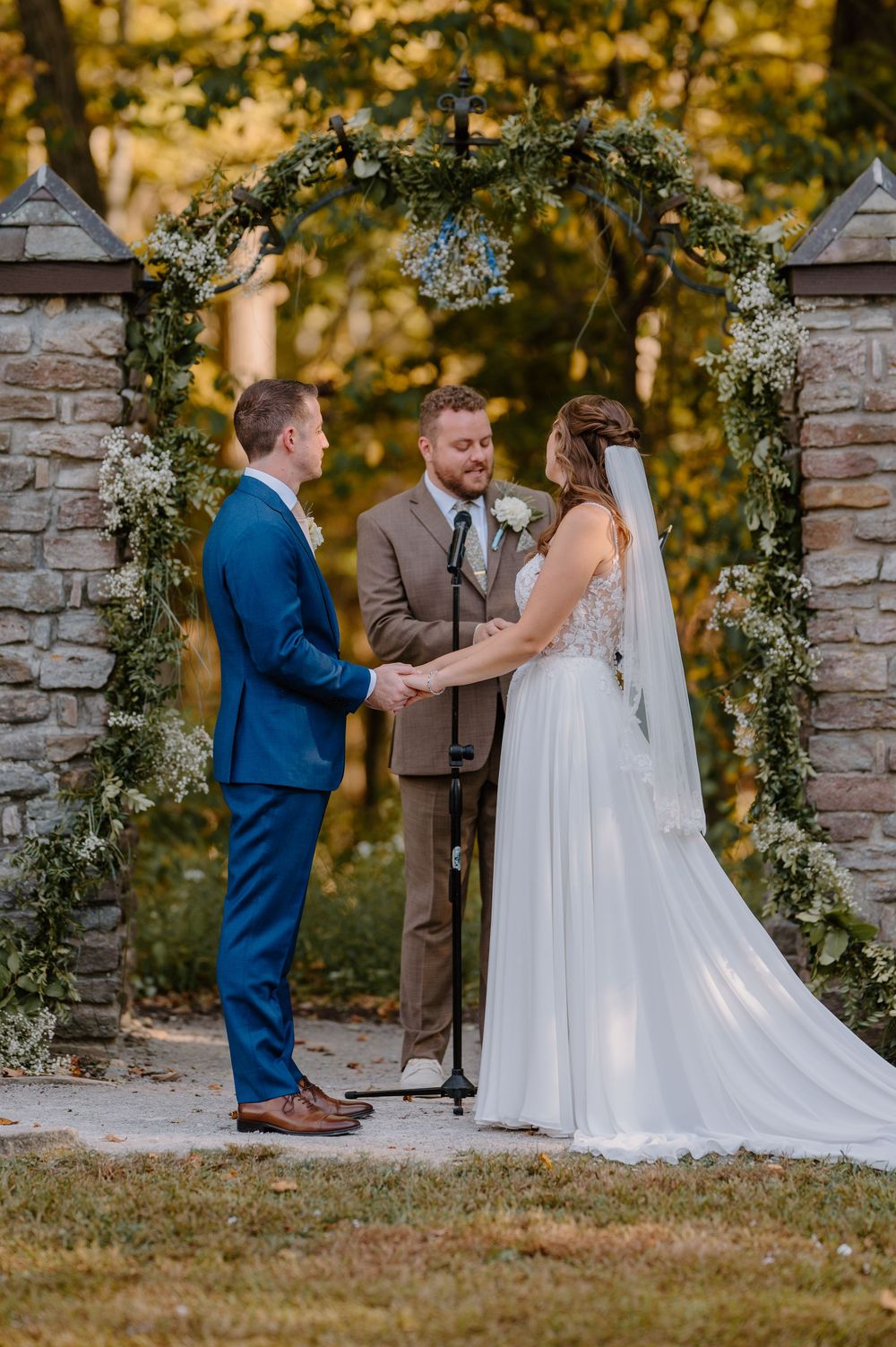 Wedding ceremony takes place under floral arch with stone pillars in golden autumn light at outdoor venue.