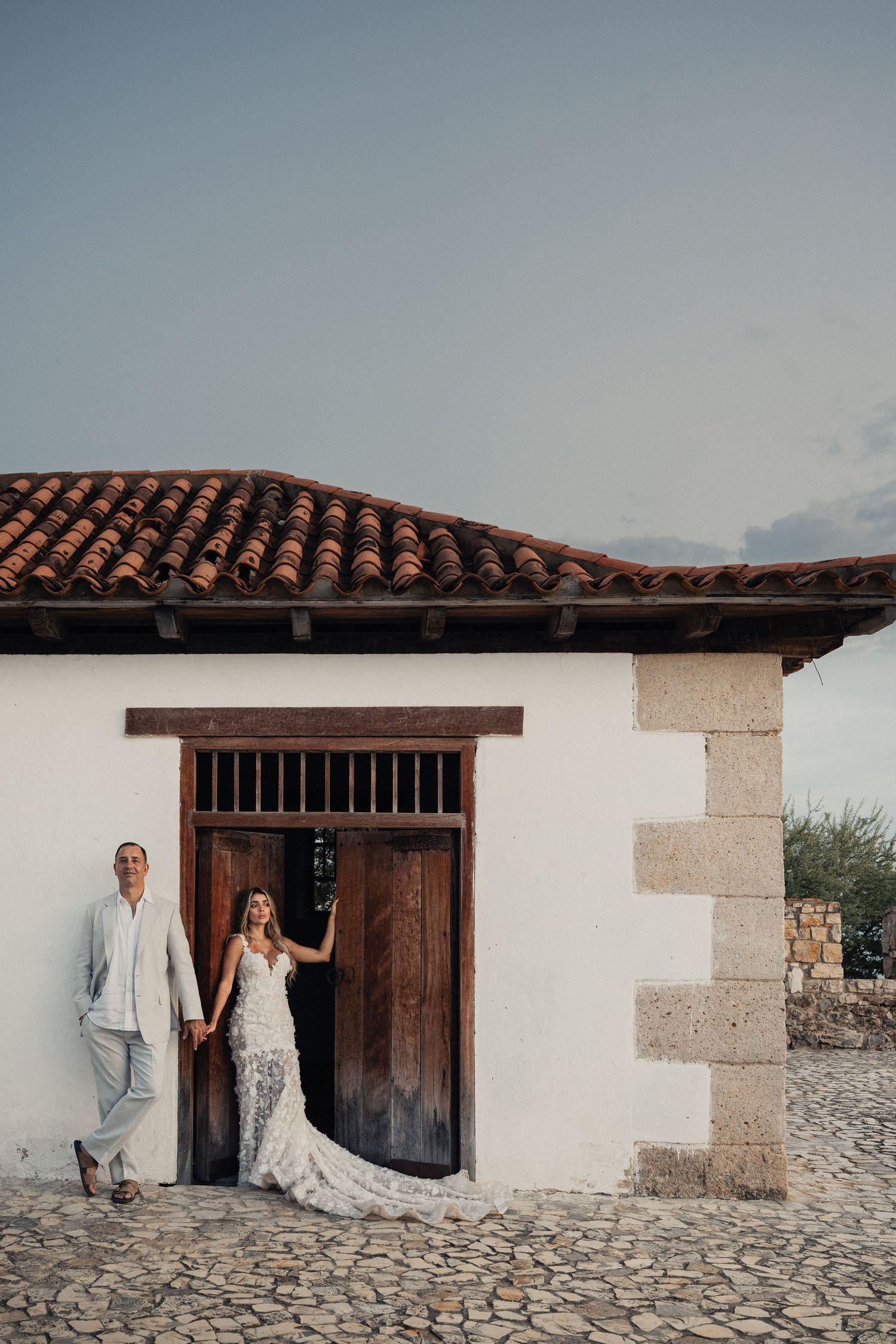 A couple stands in front of a rustic wooden door at a Mediterranean-style building with terracotta roof tiles.