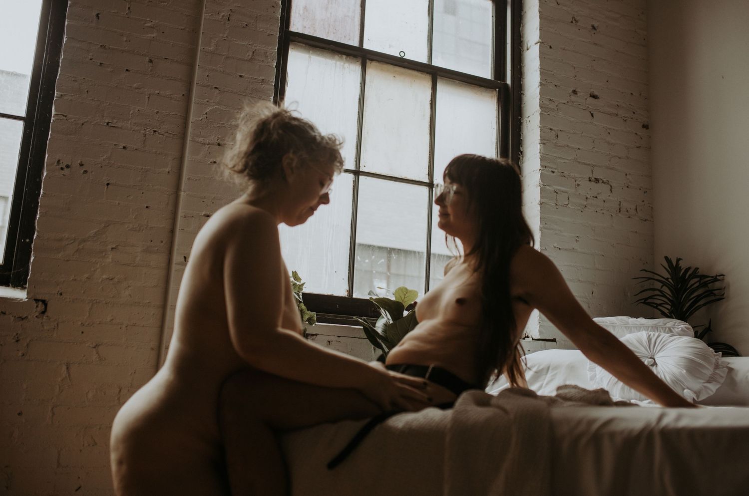 A boudoir photo showcasing the silhouettes of two queer women; one sitting on a white bed and one kneeling in front of her with moody light.