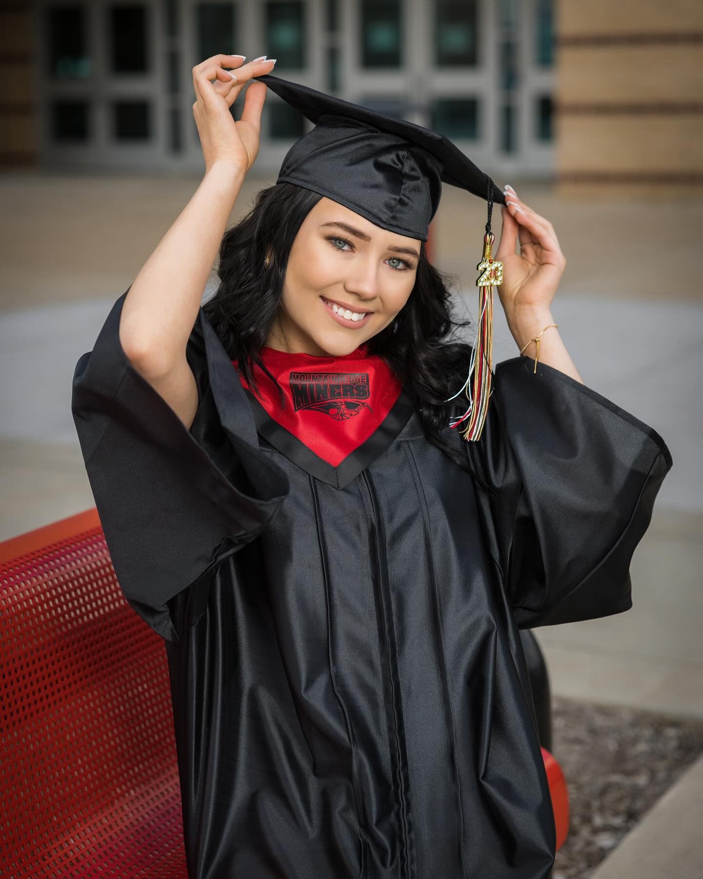 Graduate in black cap and gown with red stole adjusts their mortarboard tassel during graduation photo session.