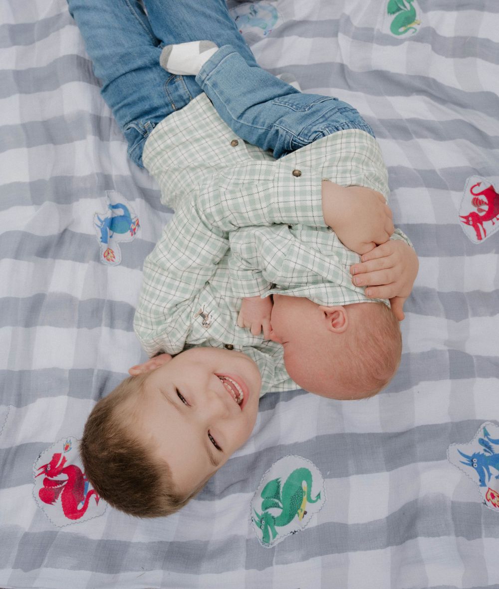 A smiling toddler boy lies on a plaid blanket with sea creature prints, holding a newborn baby sibling.