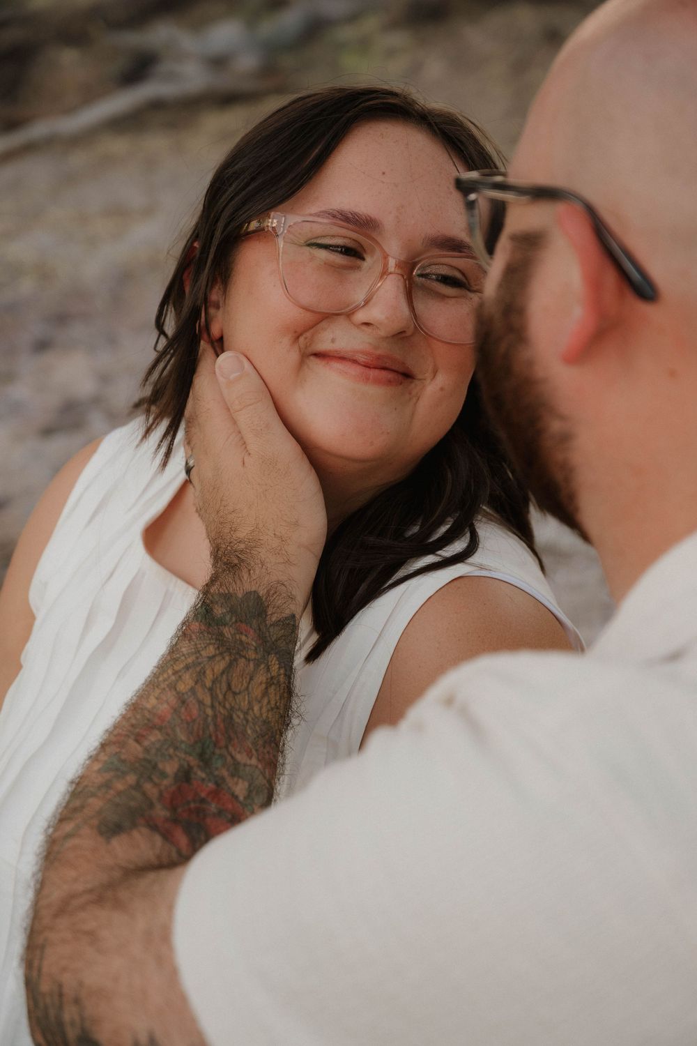 Romantic close-up of tattooed man gently cupping smiling woman with glasses face outdoors.