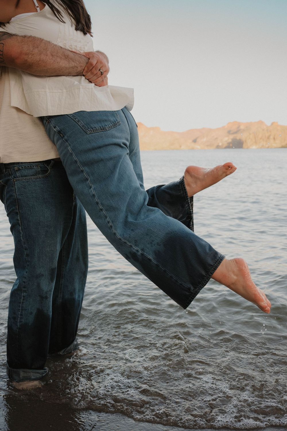 Couple embracing in shallow water at lake, woman lifted off ground, mountains in background.