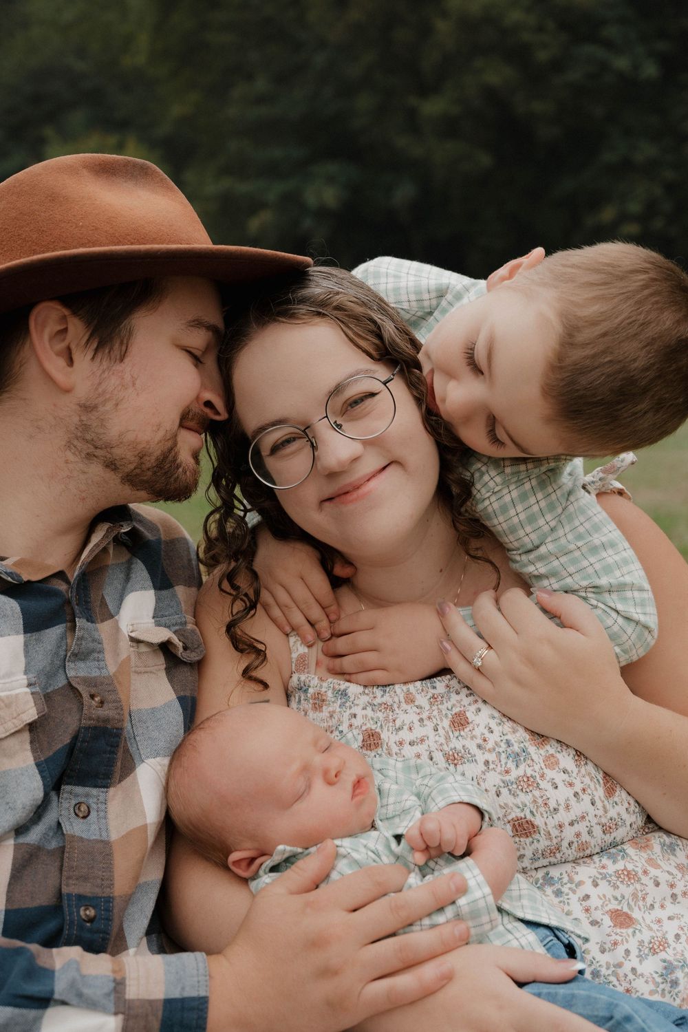 A young family of four shares a tender outdoor moment, with parents and toddler cradling their newborn baby.