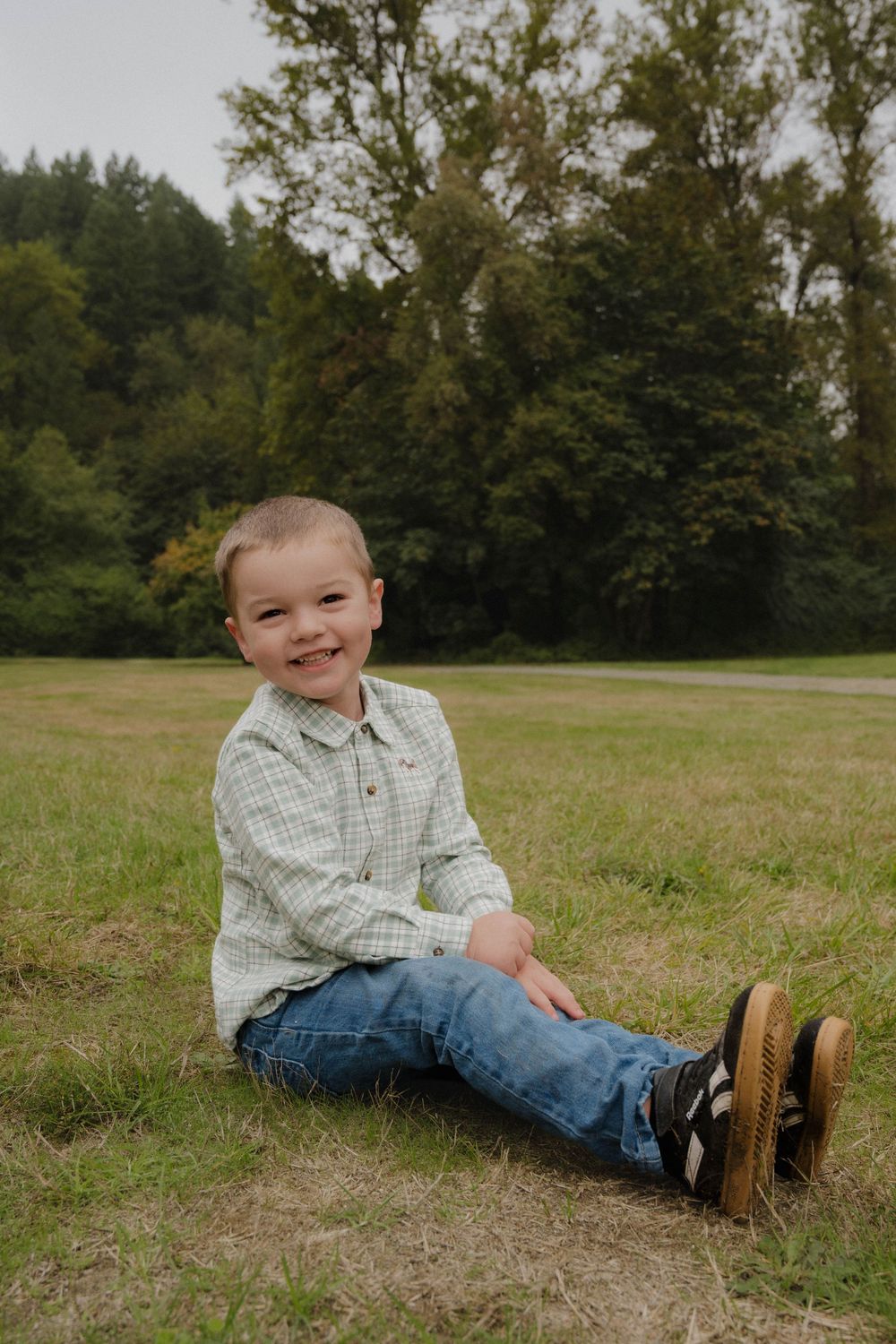A cheerful toddler boy sits on green grass in a plaid shirt and jeans, smiling outdoors near lush trees.
