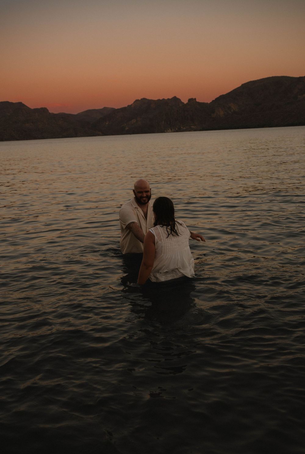 Two people stand waist-deep in calm lake water during a golden sunset baptism ceremony with mountains in the background.