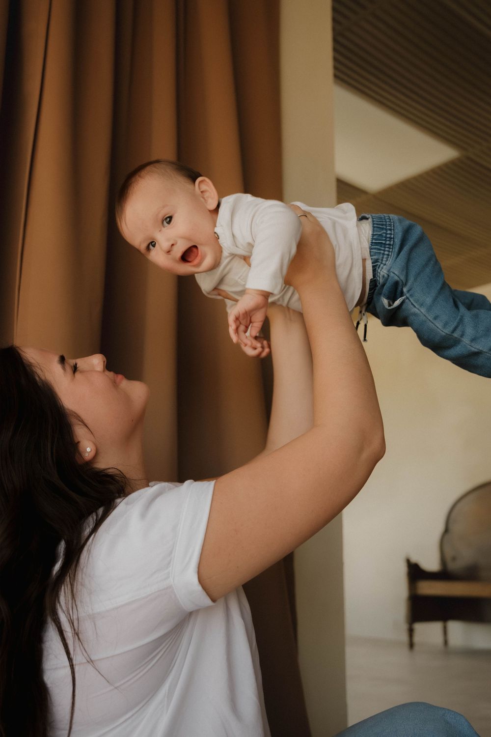 Mother in white t-shirt joyfully lifting laughing baby in white onesie and jeans indoors.