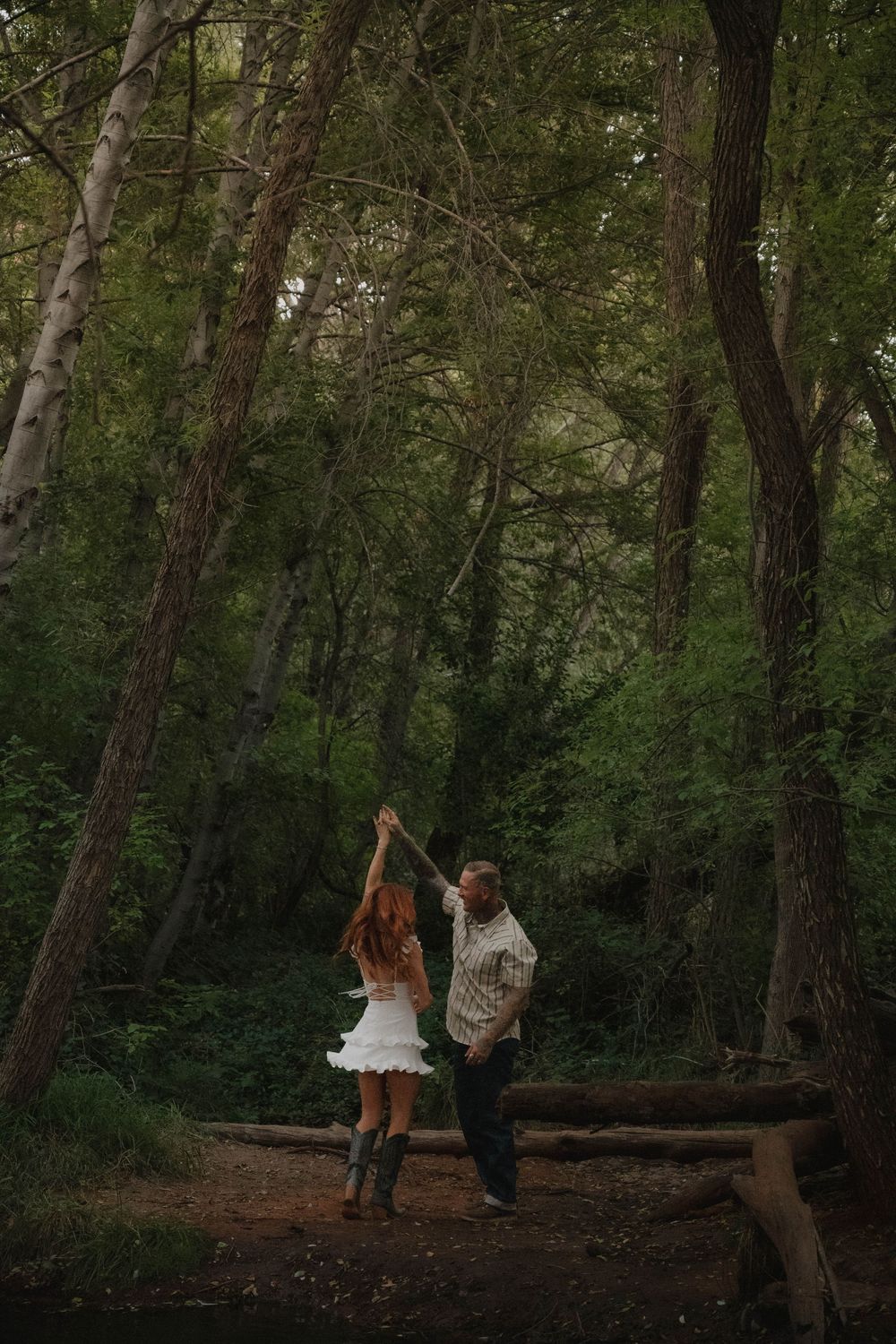 Couple dancing together in a lush green forest, woman in white dress and cowboy boots, man in casual shirt.