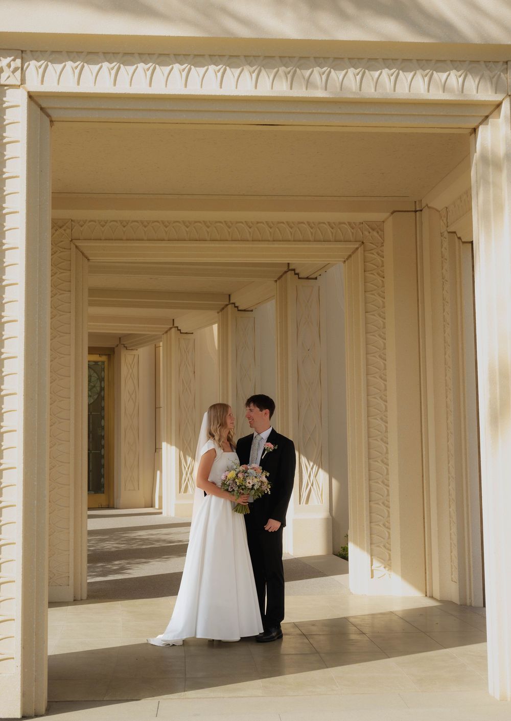Bride and groom share an intimate moment beneath elegant arched columns at an LDS temple wedding.