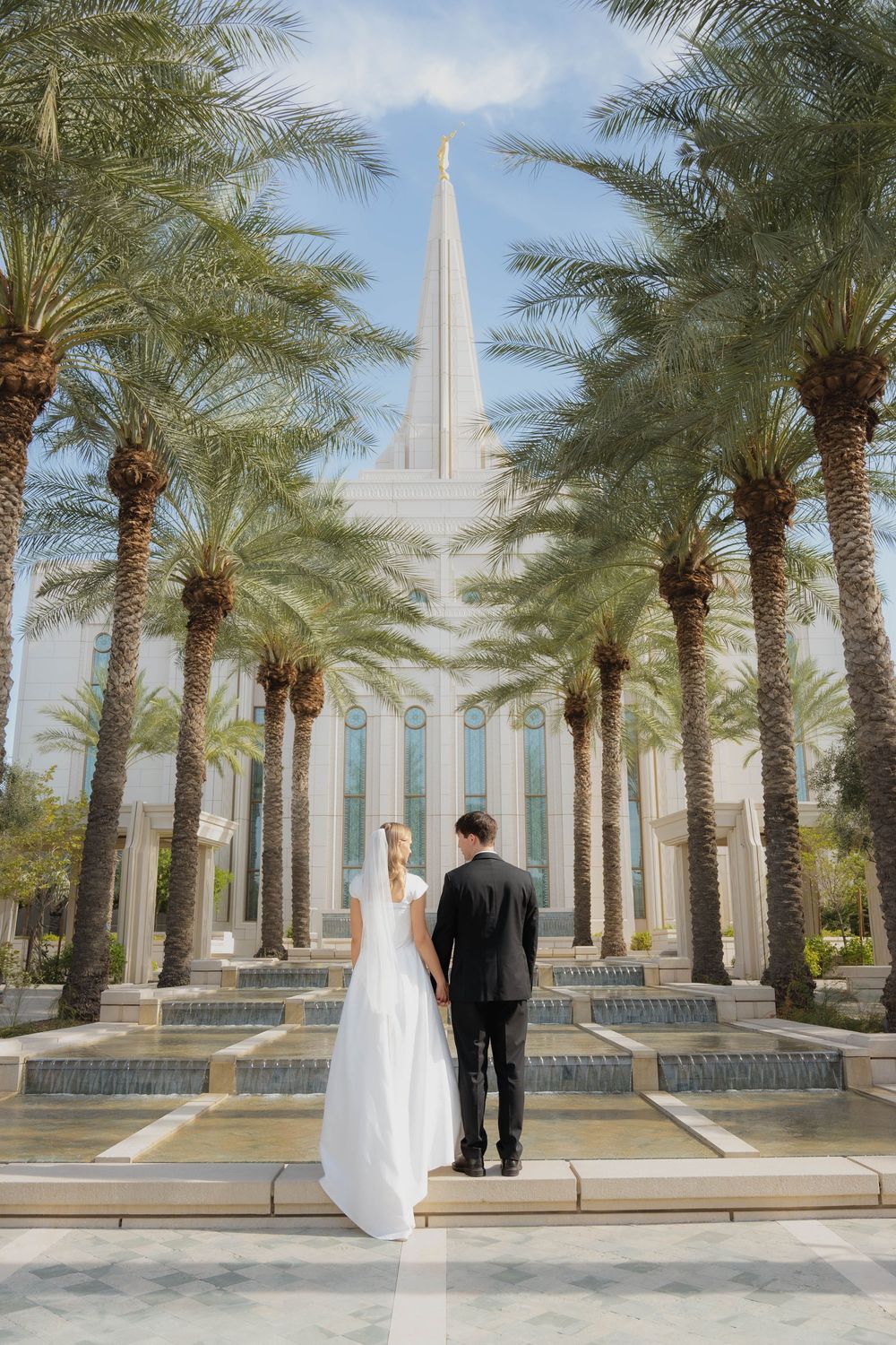 Bride and groom hold hands facing the Gilbert Arizona LDS Temple surrounded by tall palm trees.