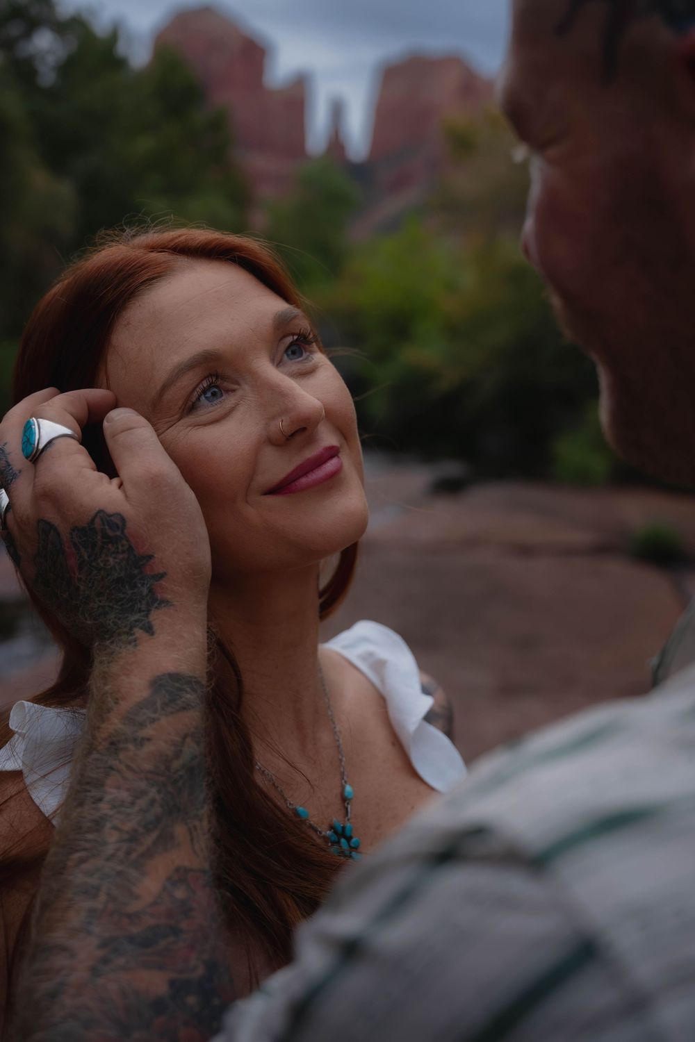 A tattooed man tenderly touches a smiling redhead woman's face near Sedona's red rock formations.