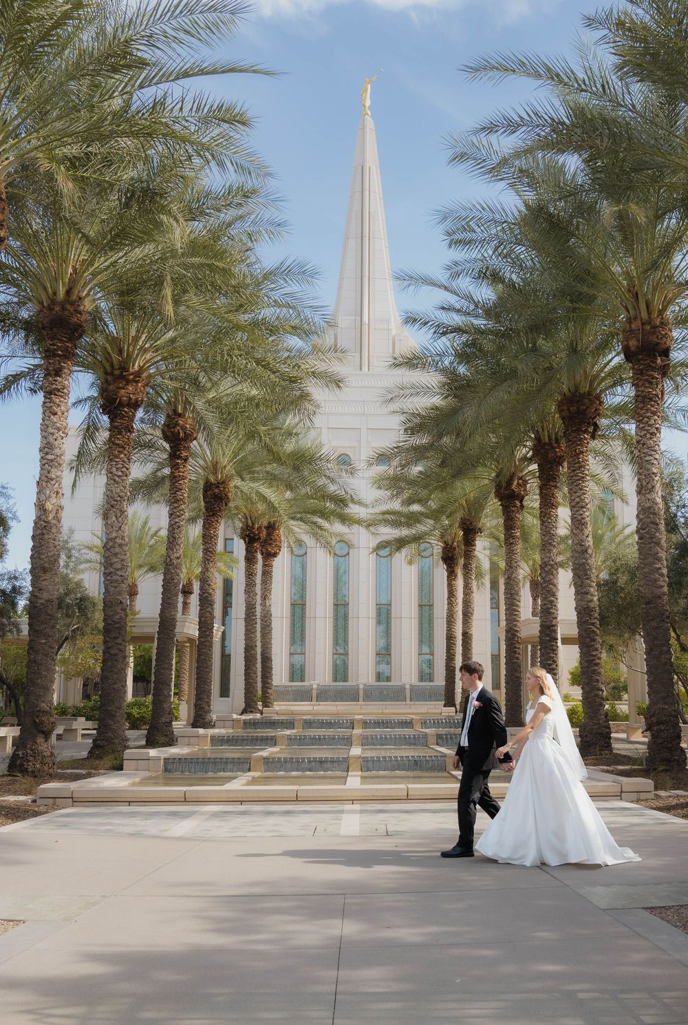 Bride and groom walk hand in hand along a palm tree-lined path toward an LDS temple spire.