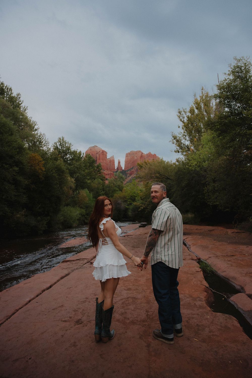 Couple holding hands on red rocks in Sedona, Arizona, with Cathedral Rock and lush green trees in the background.