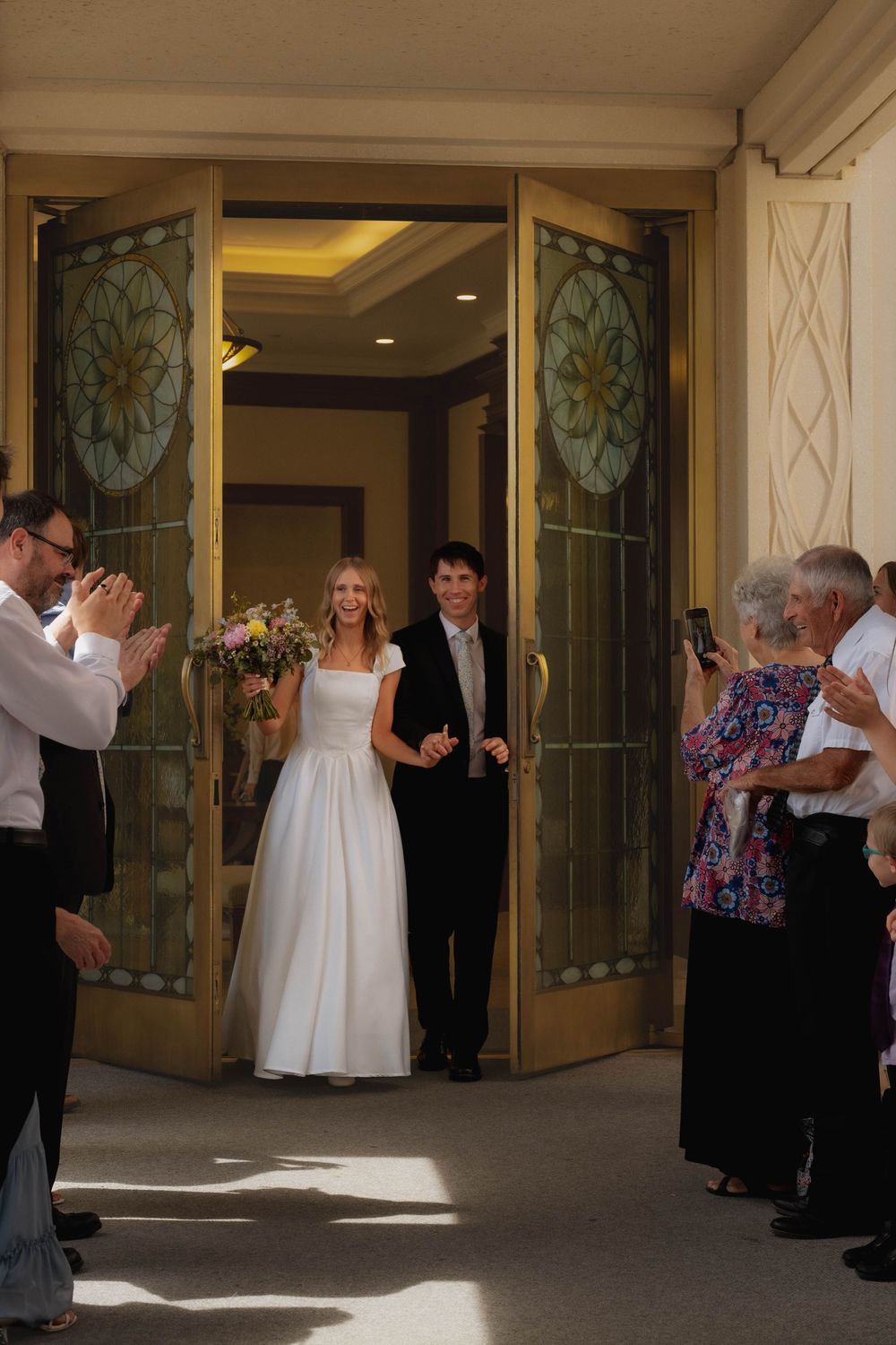Newlywed couple exits through ornate temple doors as guests applaud and celebrate their wedding.