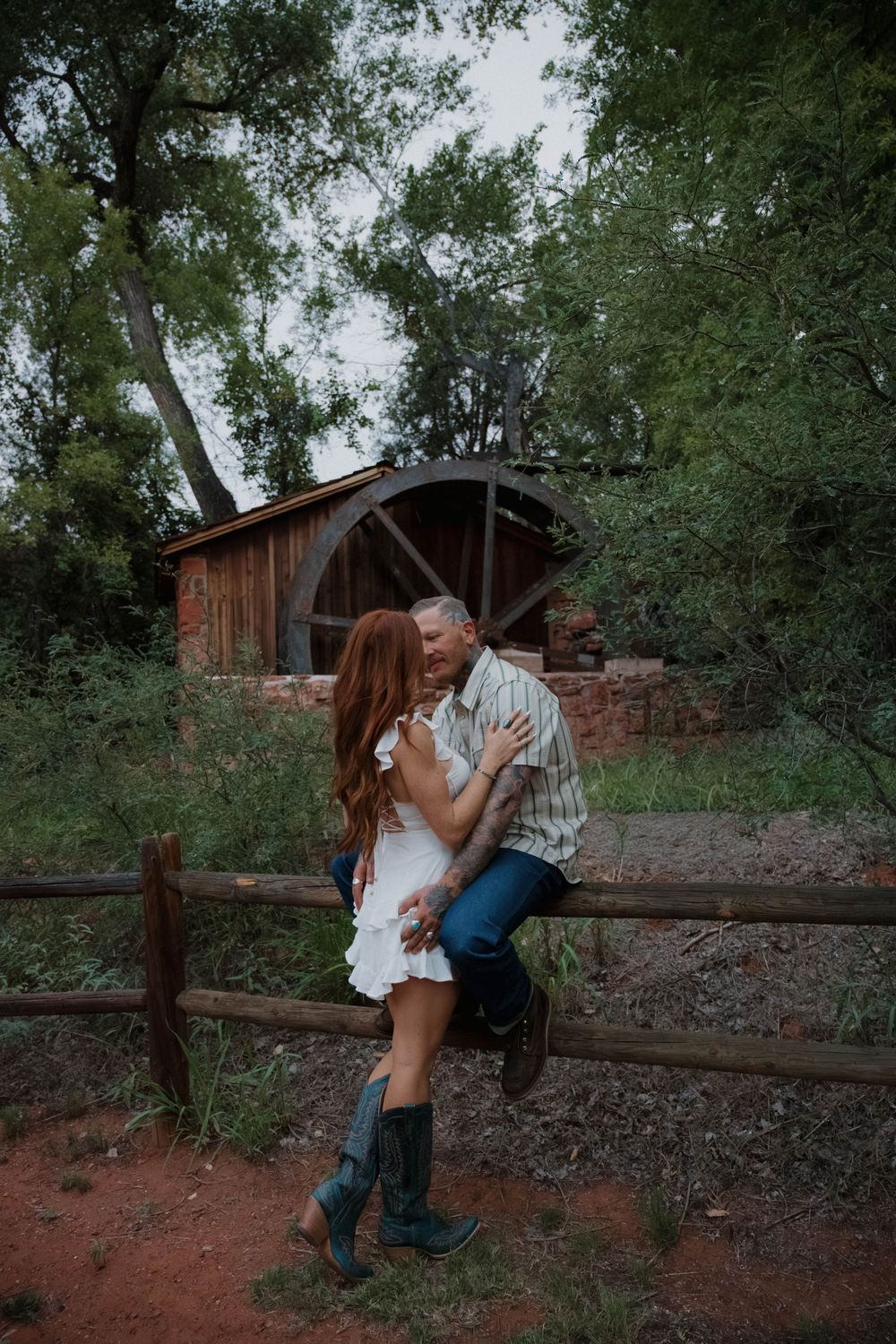 Couple embracing by a rustic wooden fence near an old water wheel surrounded by lush green trees.
