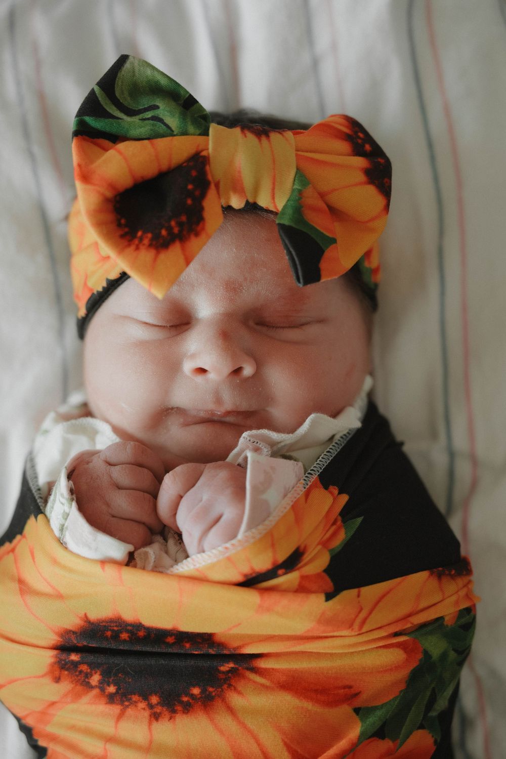 Sleeping newborn baby swaddled in a sunflower print blanket with matching floral bow headband.