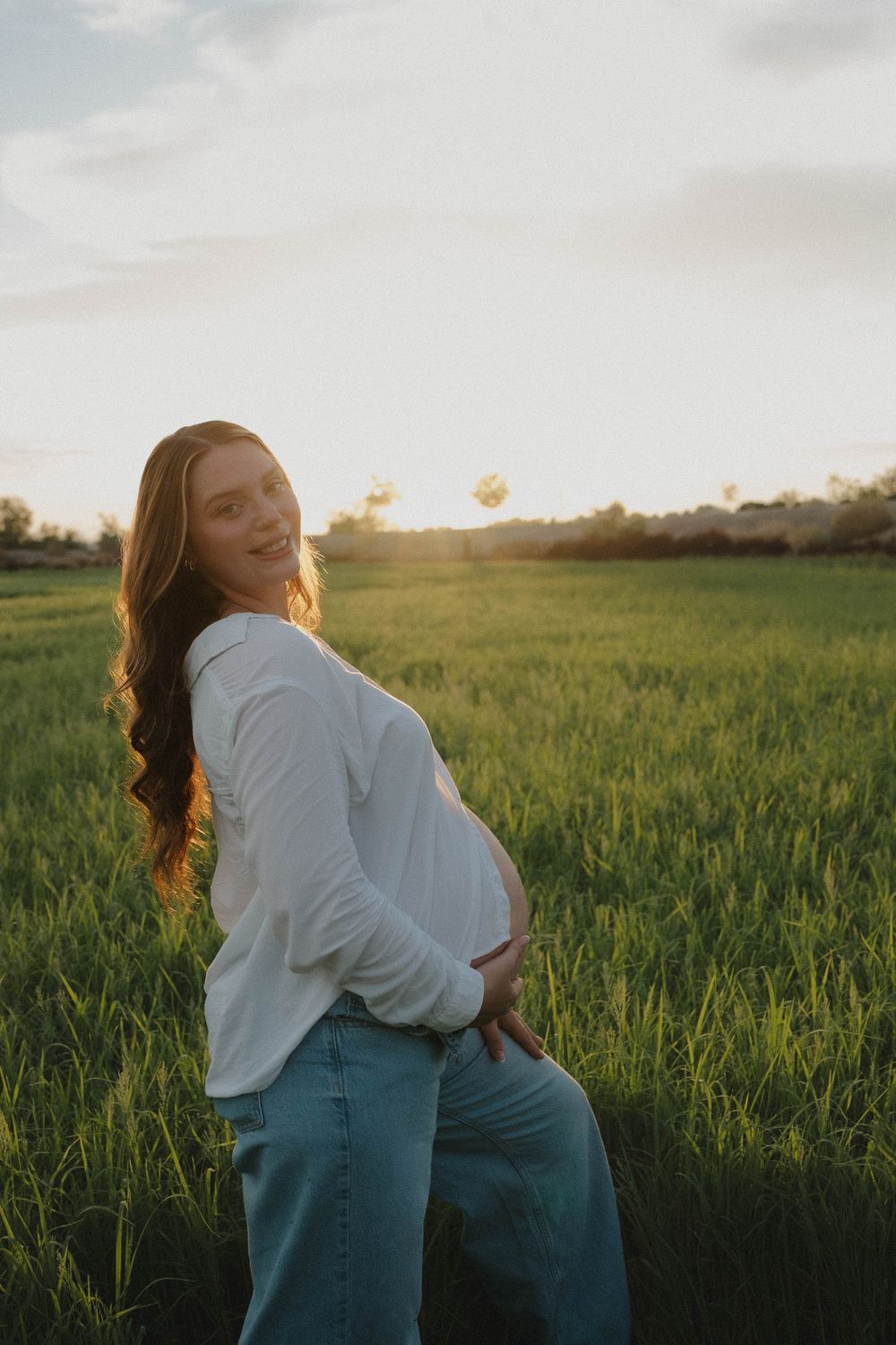 Smiling pregnant woman in white shirt and jeans cradling her baby bump in a green field at golden hour sunset.