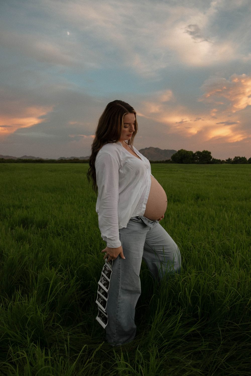 Pregnant woman in white shirt and jeans holds ultrasound photos in a green field at sunset.