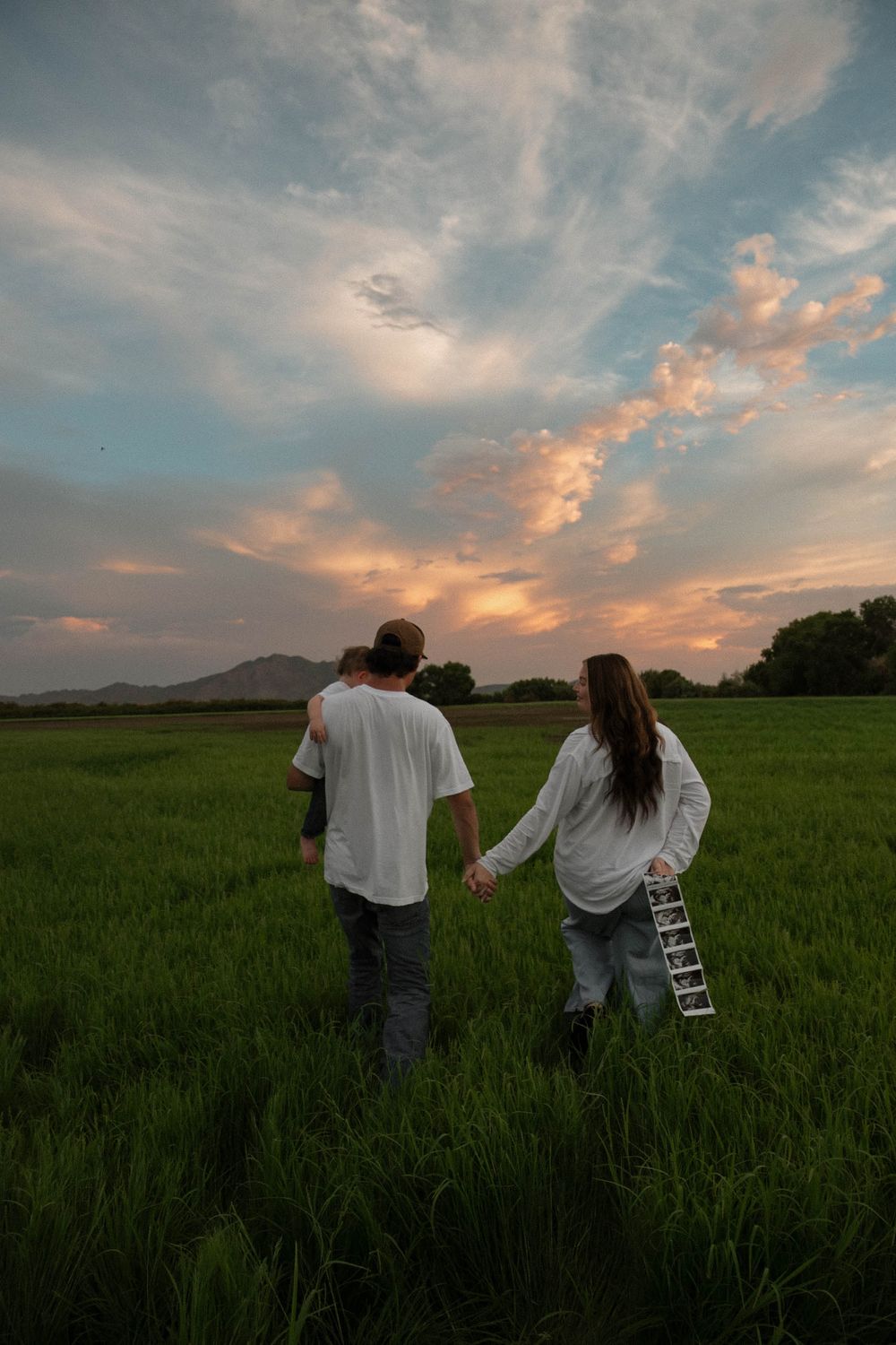 Couple holding hands walking through lush green field at sunset with dramatic cloudy sky and mountains in background.