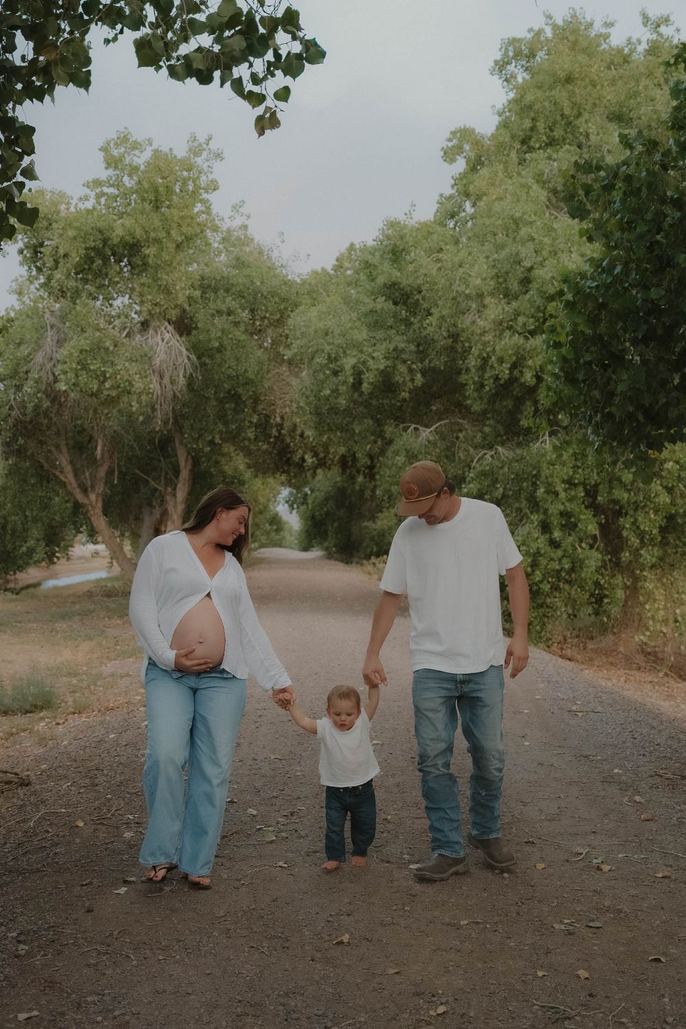 Pregnant couple holding toddler's hands on a tree-lined path during an outdoor maternity photoshoot.