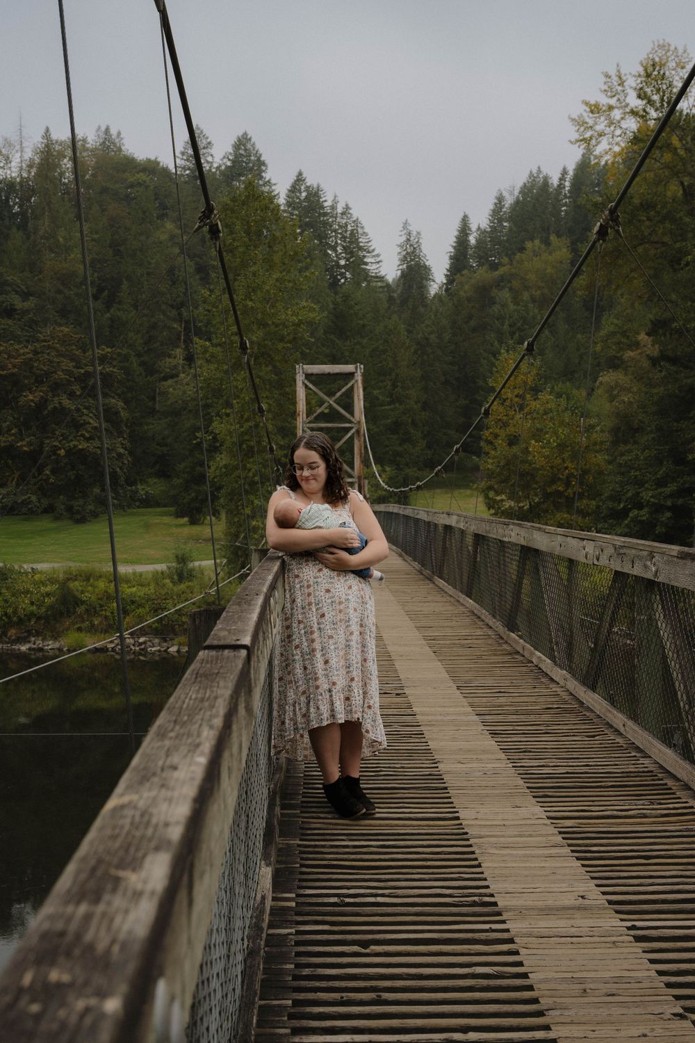 Young woman in floral dress standing on rustic wooden suspension bridge surrounded by lush green forest.