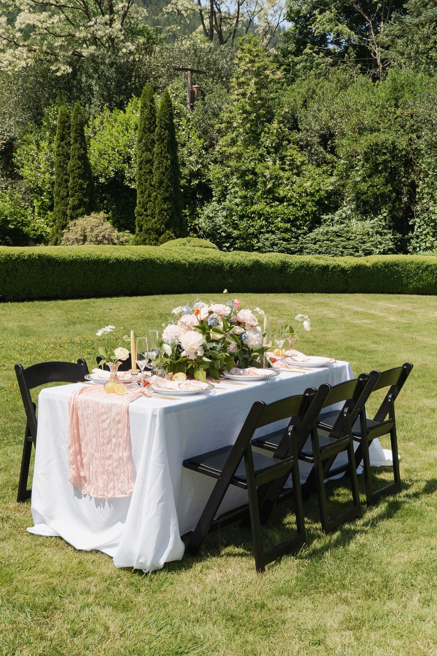 Elegant outdoor garden party table settings with white linens and pink accents arranged on a sunny lawn with trees in background.