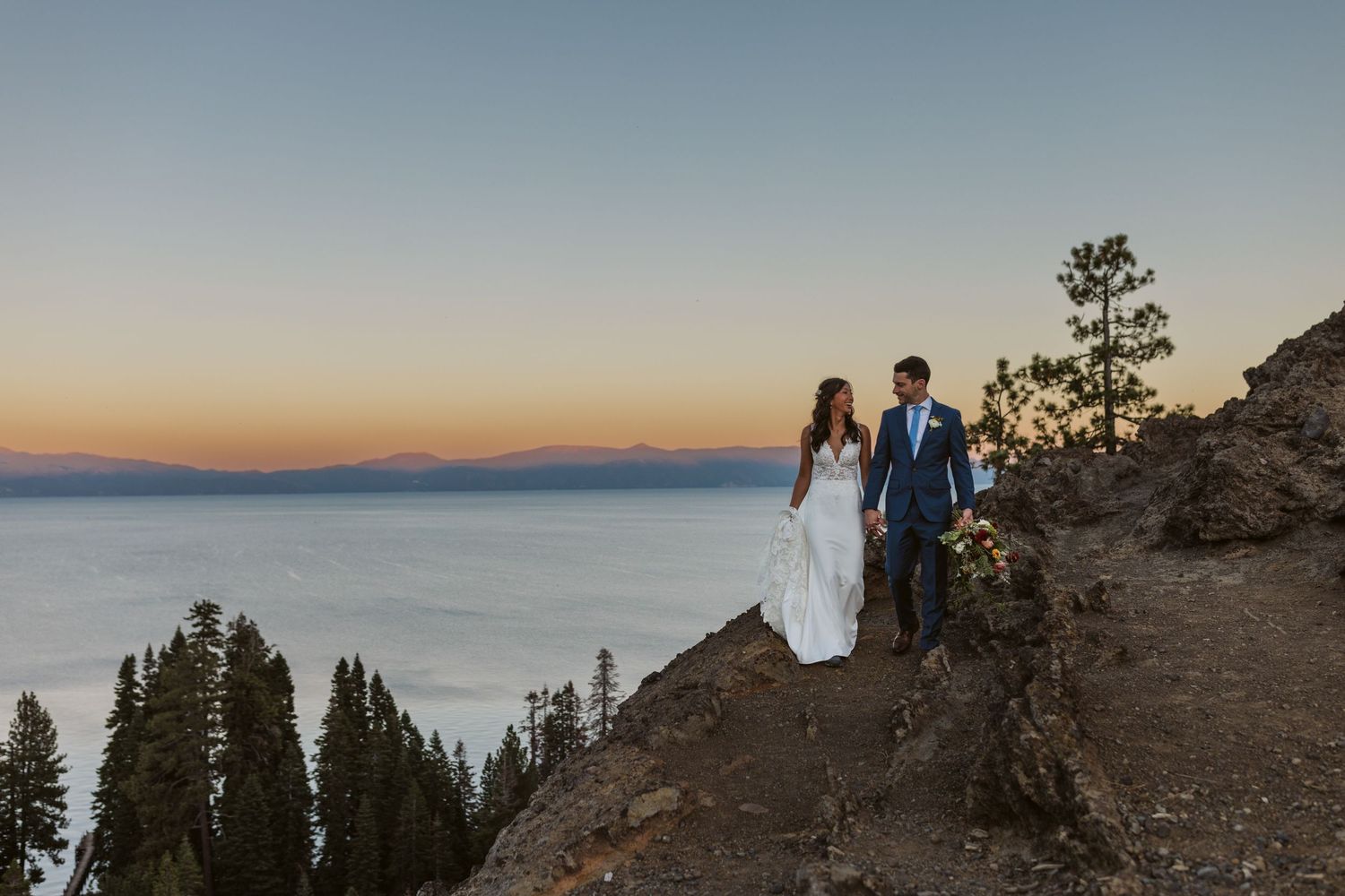 People stand on a mountain ridge silhouetted against a vibrant sunset sky with distant peaks in view.