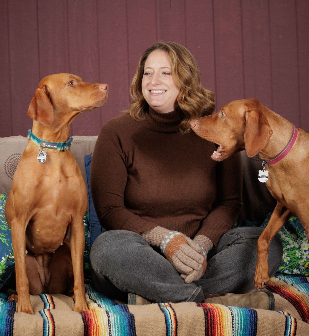 A smiling woman sits cross-legged on a couch between two golden Vizsla dogs indoors.