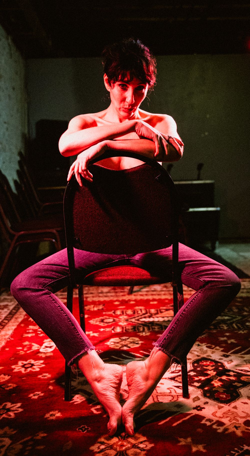 A woman sits on a chair in red light, barefoot in jeans, arms crossed over her knees.