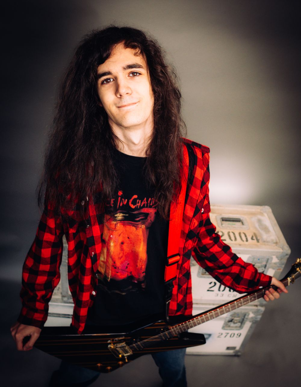 A young guitarist with long dark hair holds a guitar and cash in a red flannel shirt.